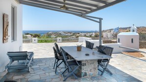 A covered outdoor seating area with a table and chairs, a view of the sea, and a white building with a cross in the background. - Villa Fotis Country House with Sea View, Wi-Fi, and Air Conditioning (Megas Gialos)