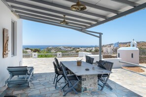 A covered outdoor seating area with a table and chairs, a view of the sea, and a white building with a cross in the background. - Villa Fotis Country House with Sea View, Wi-Fi, and Air Conditioning (Megas Gialos)
