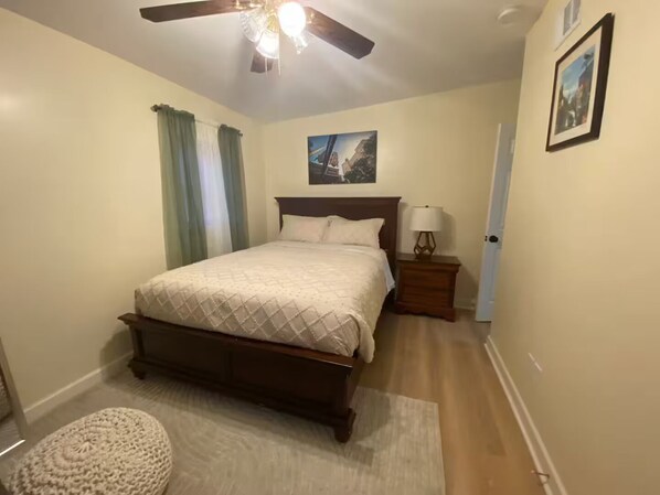 A bedroom with a bed, a nightstand, a lamp, a ceiling fan, a painting on the wall, and a door. - Joyland Jewel with Fenced in backyard (Atlanta)