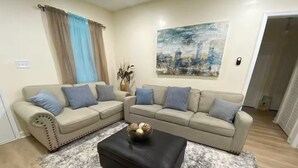 Una sala de estar con un sofá seccional color beige, un reposapiés negro, un cuadro de un paisaje urbano y una ventana con cortinas azules.
