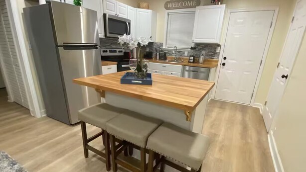 Una cocina con una isla de madera, banco, refrigerador de acero inoxidable, microondas y armarios blancos.