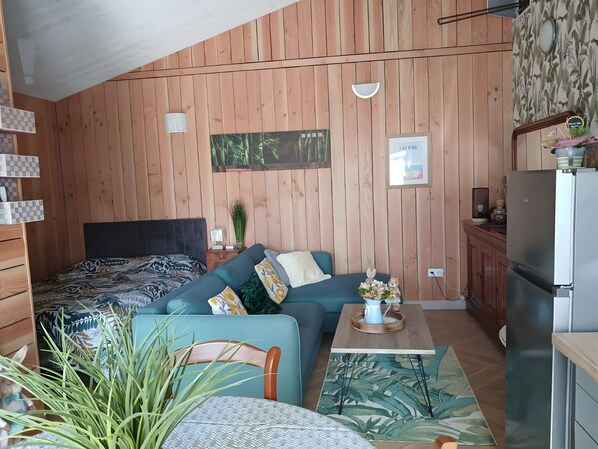 A room with wooden walls, a bed, a sofa, a dining table, a chair, a refrigerator, and a small kitchen area.