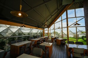Un restaurante con una decoración moderna, que incluye un cerramiento de vidrio, mesas de madera y lámparas colgantes.