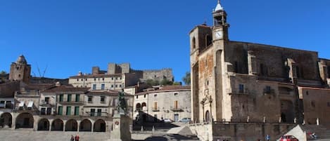 Una plaza histórica con una imponente torre del reloj, edificios de piedra y una estatua.