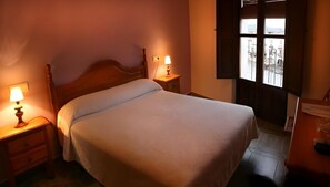 A bedroom with a bed, two bedside lamps, a nightstand, a dresser, and a window with a view of buildings.