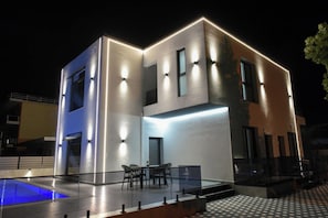 Un edificio moderno de dos plantas con piscina, mobiliario exterior y fachada bien iluminada.