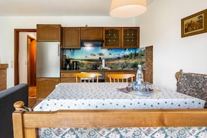 Un coin repas avec une table recouverte d’une nappe à motifs étoilés, des chaises en bois et, au fond, une cuisine avec des meubles en bois et une fresque murale.