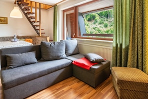 Ein modernes Wohnzimmer mit einem grauen Ecksofa, einem kleinen Hocker, einem Esstisch mit einer Wasserflasche, einer Treppe und einem Fenster mit Vorhängen.