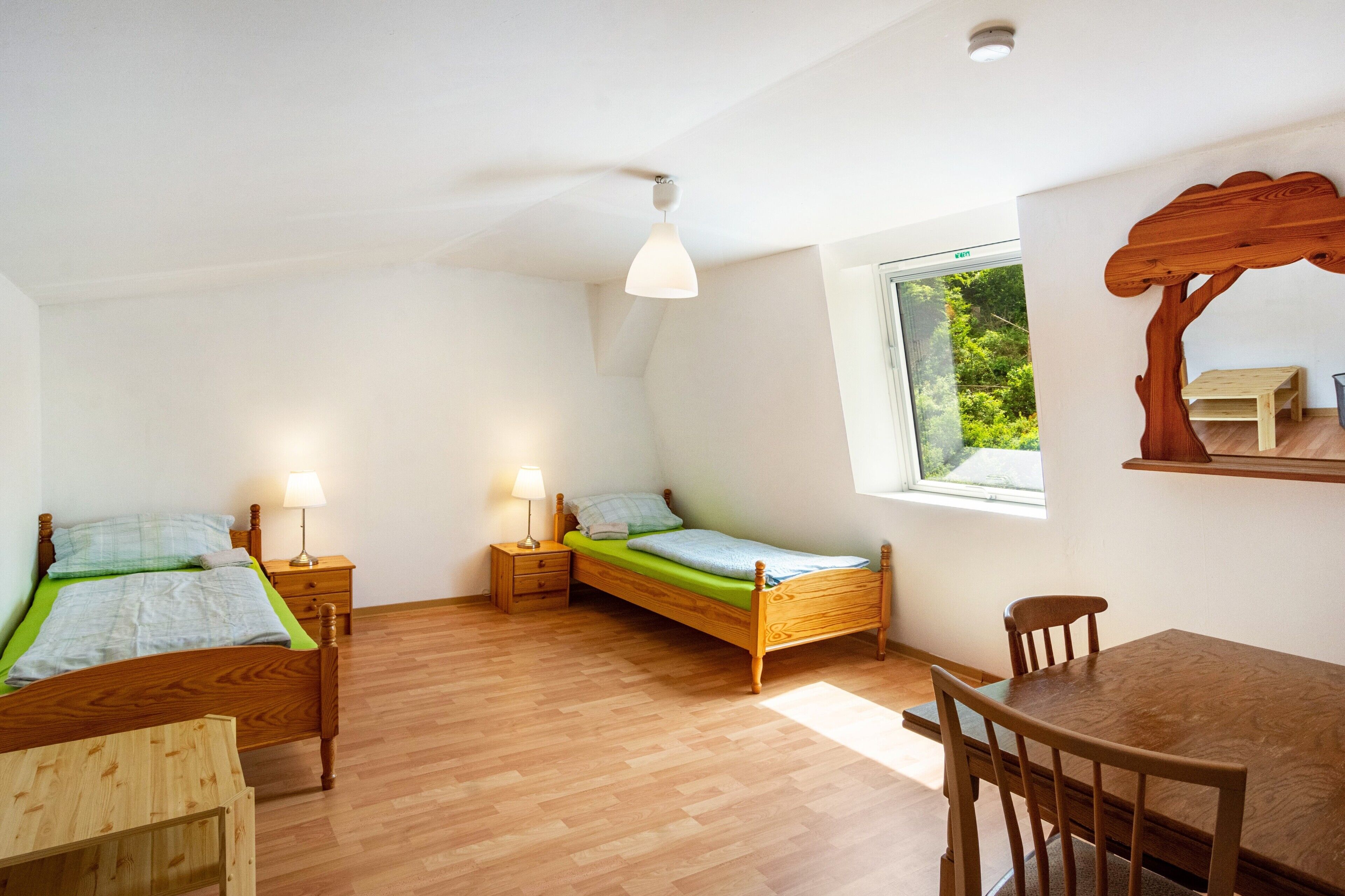 A room with two wooden beds, a wooden table, and a window with a view of greenery.