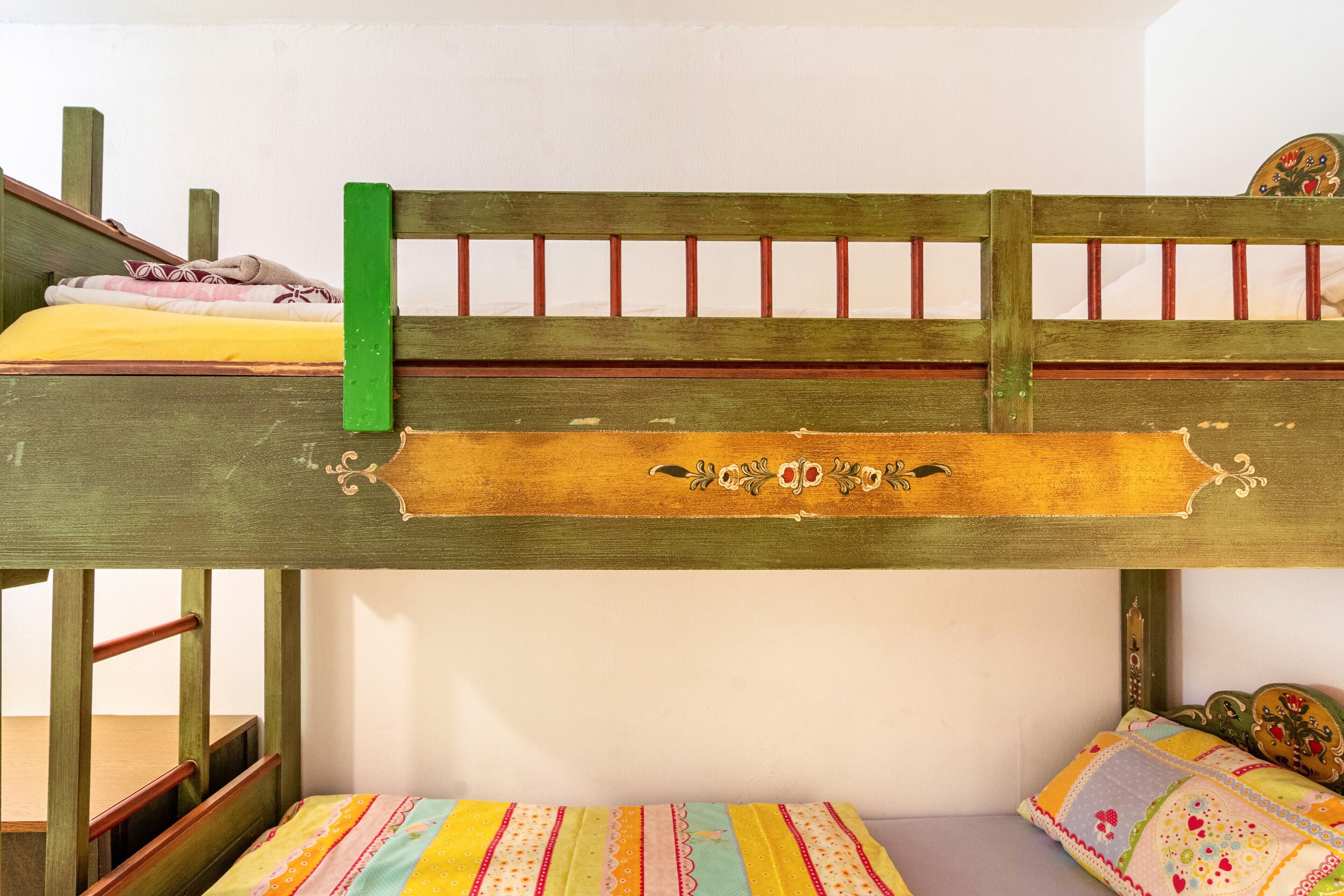 A bunk bed with a green headboard, a lower bed with a colorful quilt, and a wooden chair.