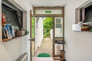 Ein schmaler Flur mit einer Tür, auf der 'Ausgang' steht, die zu einem Garten führt. Links befinden sich Regale mit Gegenständen, rechts ein Heizkörper und vor der Tür eine kleine grüne Matte.