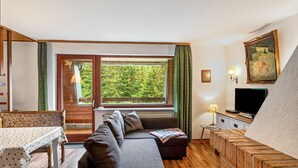Ein modernes Wohnzimmer mit einer Couch, einem Esstisch, einem Fernseher und einem großen Fenster mit Blick auf Grünflächen.