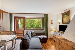 Ein modernes Wohnzimmer mit einer Couch, einem Esstisch, einem Fernseher und einem großen Fenster mit Blick auf Grünflächen.