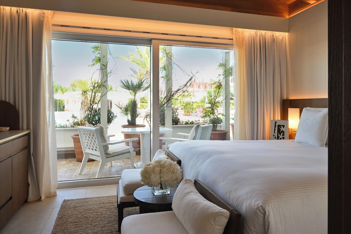 A hotel room with a large bed, a desk with a chair, a small table with flowers, and a view of the outdoor patio.