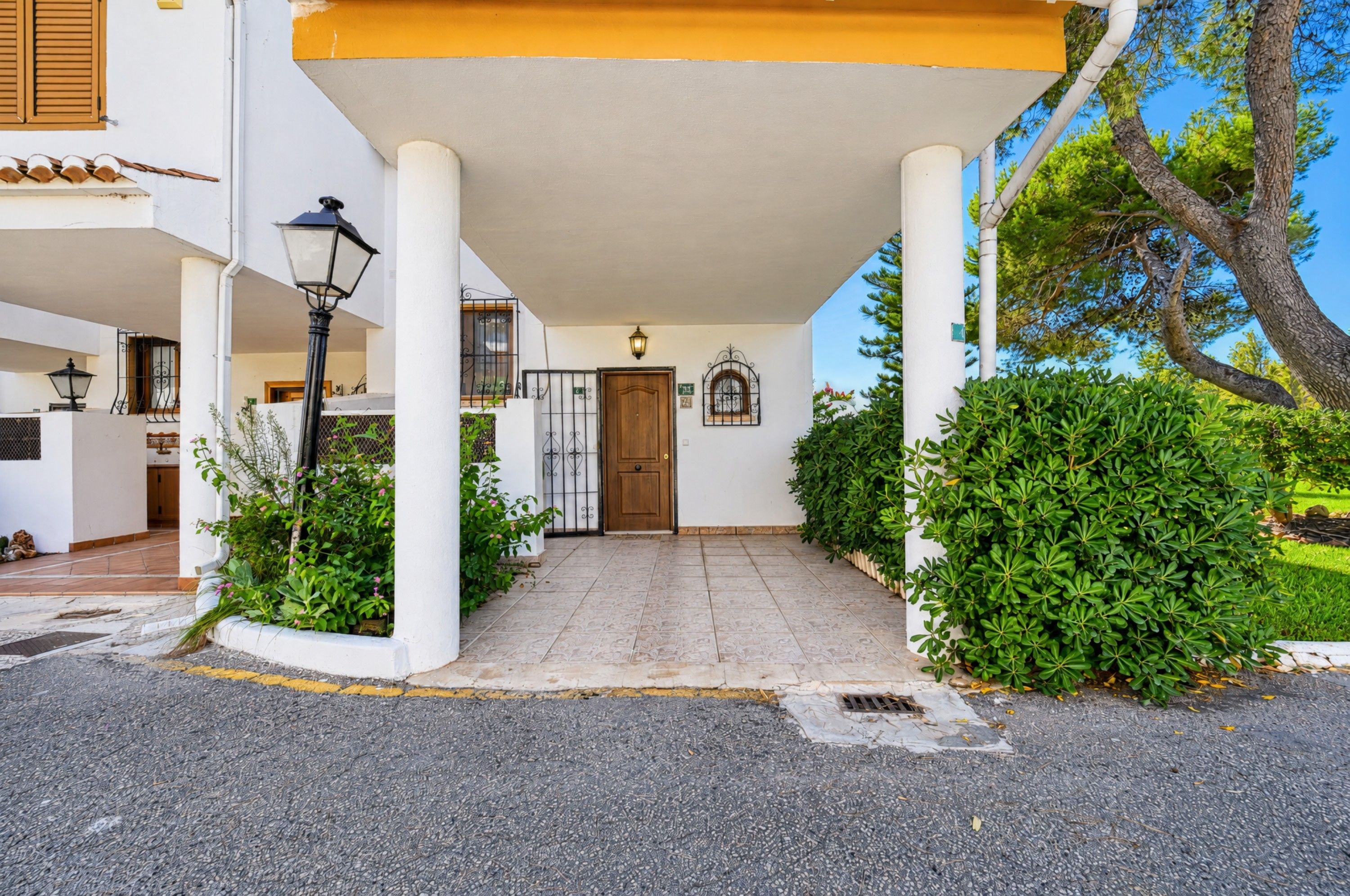 A building entrance with a tiled pathway leading to a wooden door, surrounded by greenery and a clear sky.