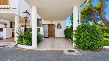 A building entrance with a tiled pathway leading to a wooden door, surrounded by greenery and a clear sky.