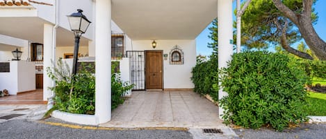 A building entrance with a tiled pathway leading to a wooden door, surrounded by greenery and a clear sky.