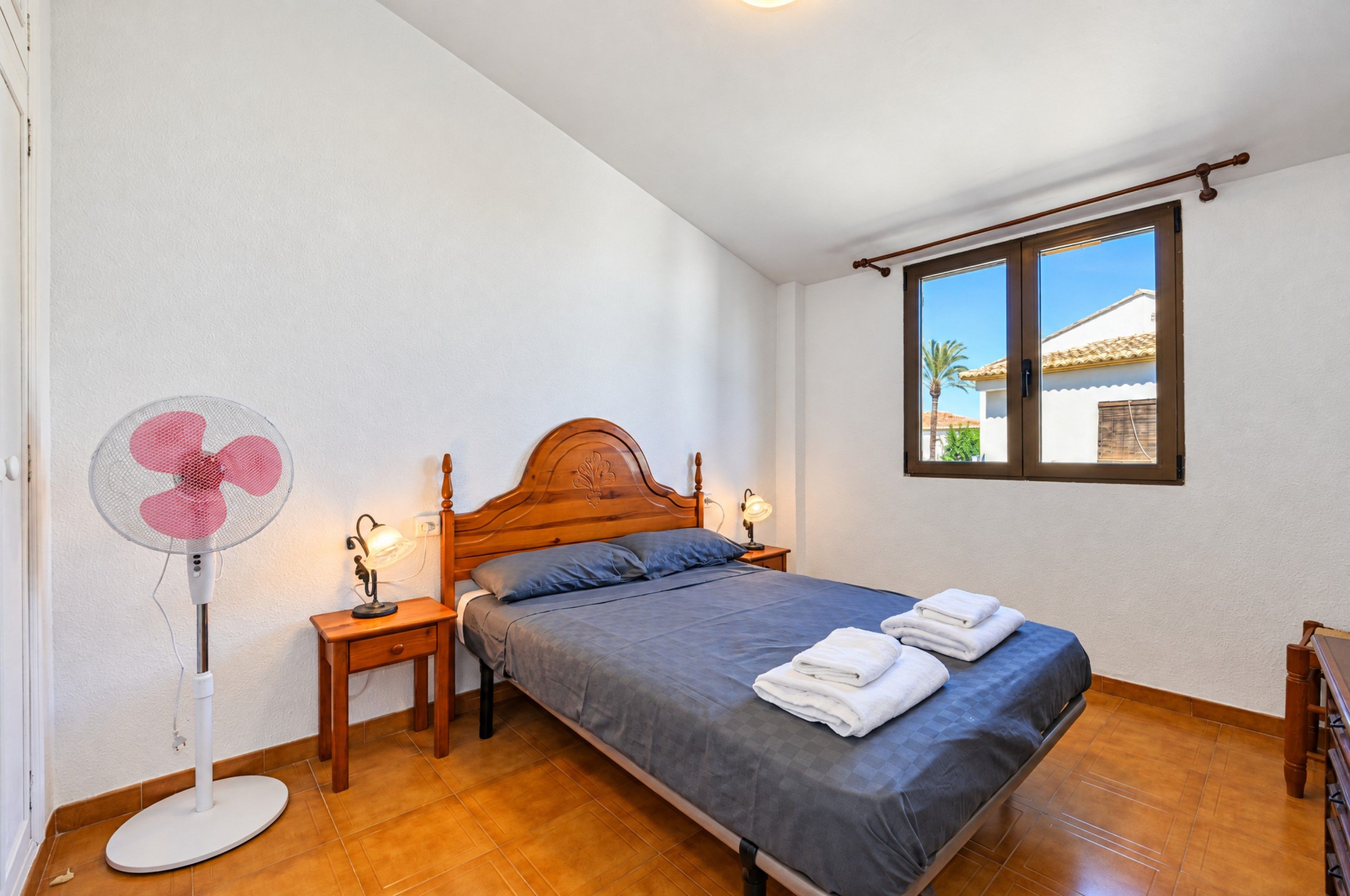 A bedroom with a single bed, two bedside tables, a standing fan, and a window with a view of a building.