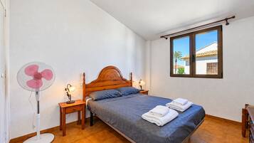 A bedroom with a single bed, two bedside tables, a standing fan, and a window with a view of a building.