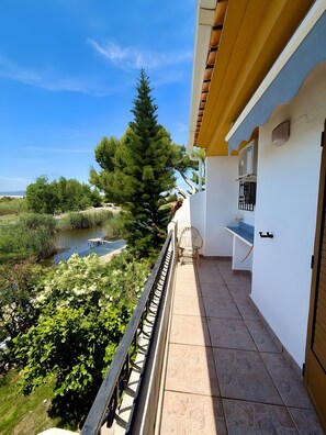 A balcony with a view of a river and trees, a white building with a yellow roof, and a small outdoor seating area.