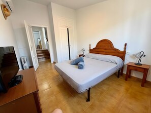 Quarto com cama de madeira, escrivaninha com computador, mesa de cabeceira com abajur e vista para uma escada que leva a outro cômodo.