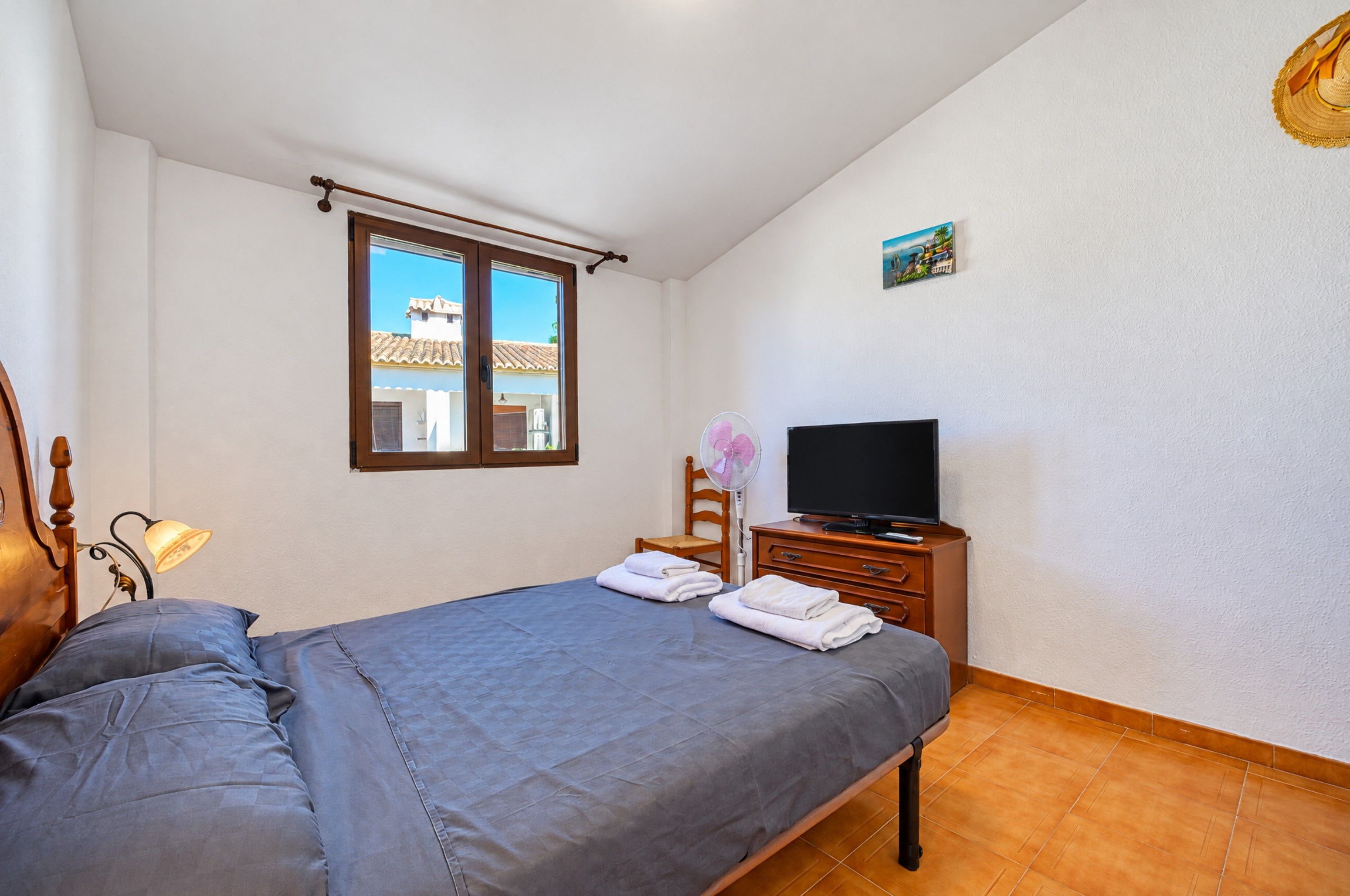 Ein Schlafzimmer mit einem Bett, einem Fernseher auf einer Holzkommode, einem Fenster mit Blick auf Gebäude und einem an der Wand hängenden Bild.