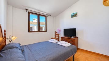 Ein Schlafzimmer mit einem Bett, einem Fernseher auf einer Holzkommode, einem Fenster mit Blick auf Gebäude und einem an der Wand hängenden Bild.