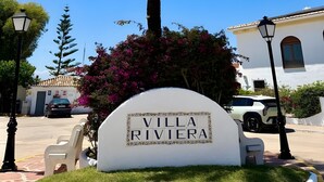 Placa de 'Villa Riviera', com uma palmeira e flores roxas ao primeiro plano.