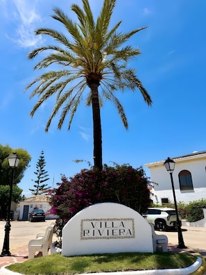 Señal de 'Villa Riviera' con una palmera y flores púrpuras al frente.