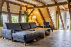 Ein Loft-Zimmer mit Ecksofa, Couchtisch und Stuhl. Sichtbare Holzbalken und große Fenster prägen den Raum.