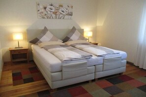 Una cama doble con una paleta de colores neutros, dos mesitas de noche con lámparas, una alfombra estampada y una obra de arte floral en la pared.