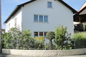 Una casa de dos pisos con fachada blanca, un balcón en el nivel superior y una cerca metálica al frente.