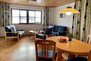 Ein Wohnzimmer mit einer Couch, Sesseln, einem Esstisch mit Obst und einem Fenster mit Vorhängen.