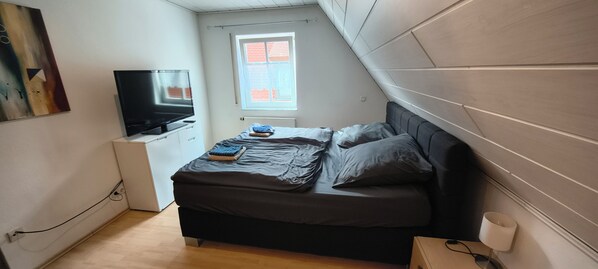 A bedroom with a bed, a television, a window, and a painting on the wall.