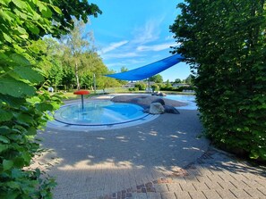 A circular pool with a blue shade sail, surrounded by greenery and paved walkways.