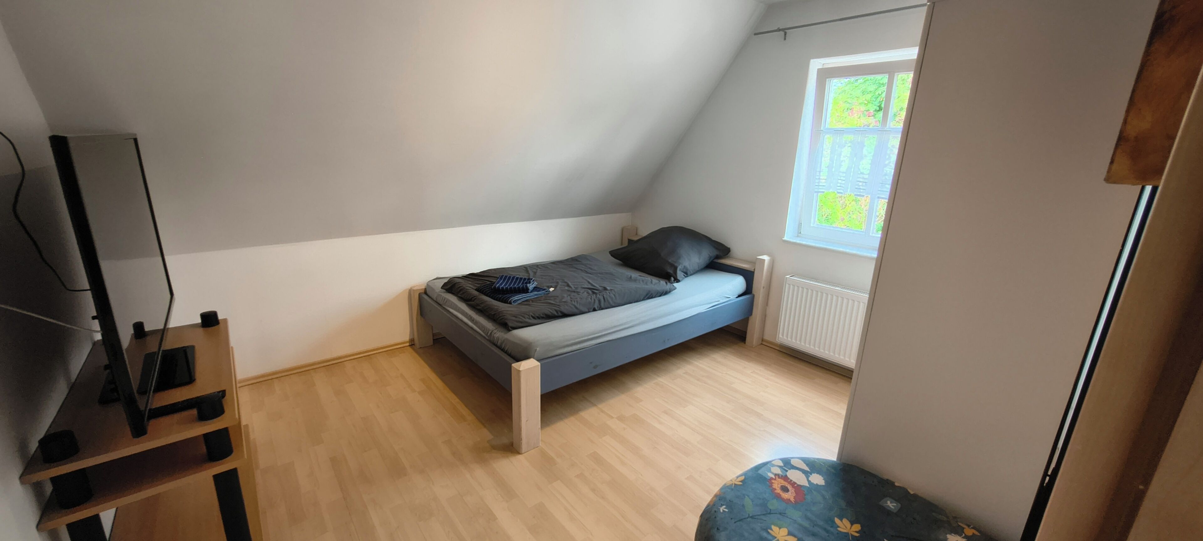 A small, single-bed room with a sloped ceiling, a window with a view of greenery, a radiator, and a TV stand.