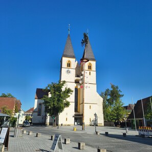 Eine Kirche mit zwei Türmen und einer Uhr, umgeben von einem gepflasterten Platz mit Bäumen und einem Schild.