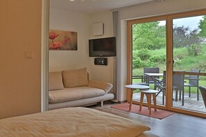 Ein Schlafzimmer mit Bett, einer Couch, einem Fernseher und einer Glastür, die auf eine Terrasse mit Stühlen und einem Tisch führt.