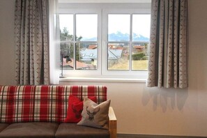 Ein Zimmer mit einer Couch, einem Fenster mit Vorhängen und Blick auf Gebäude und Berge.