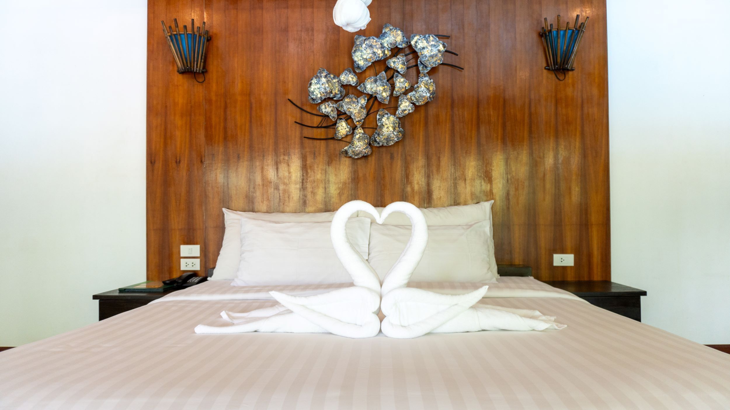 A bed with a heart-shaped towel arrangement, wooden headboard, and two wall sconces.