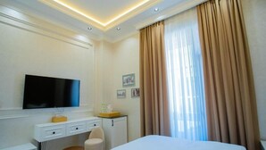 Habitación de hotel con cama, escritorio con televisor, silla, armario pequeño y ventana con cortinas.