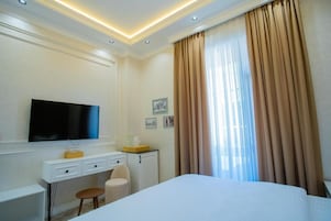 A hotel room with a bed, a desk with a TV, a chair, a small cabinet, and a window with curtains.