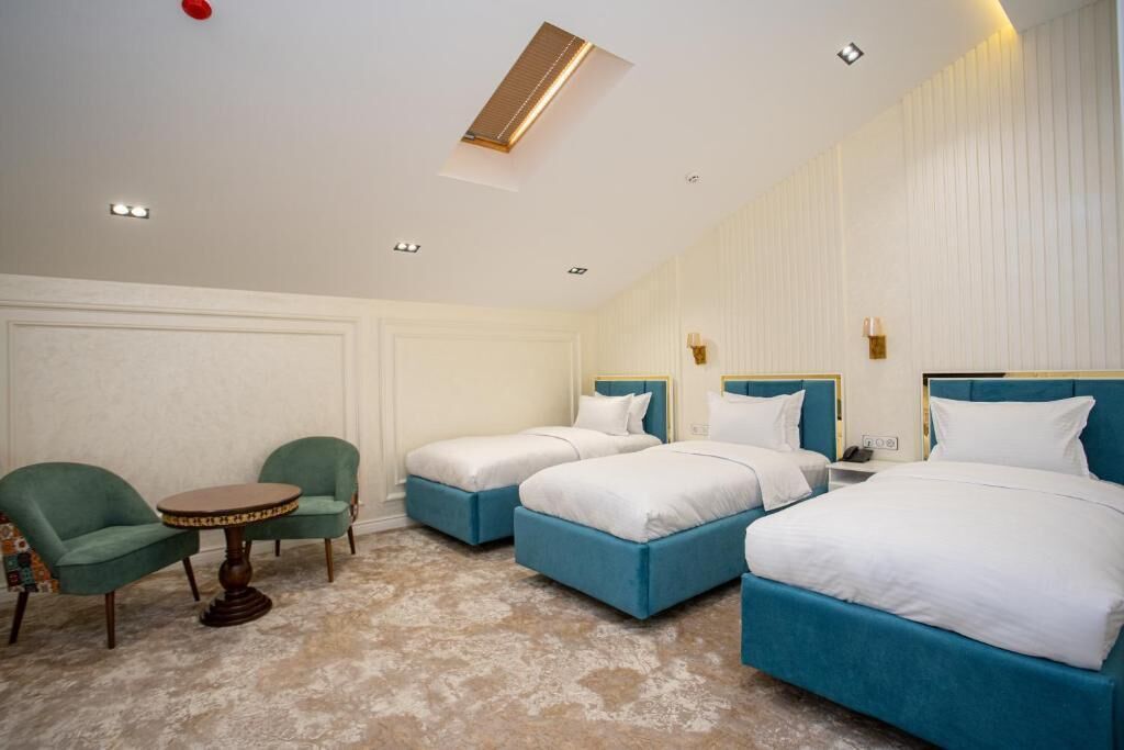 A hotel room with three beds, a small round table, two green armchairs, and a ceiling light.