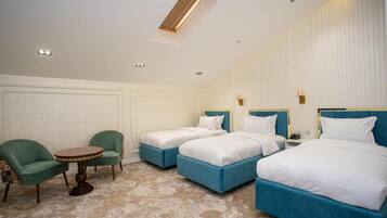 A hotel room with three beds, a small round table, two green armchairs, and a ceiling light.