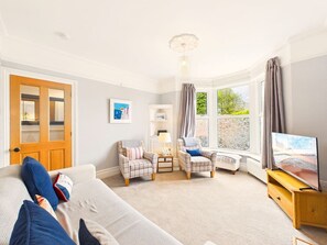 Ein Schlafzimmer mit einem Sofa, zwei Sesseln, einem Fernseher, einer Lampe und einem Fenster mit Vorhängen.
