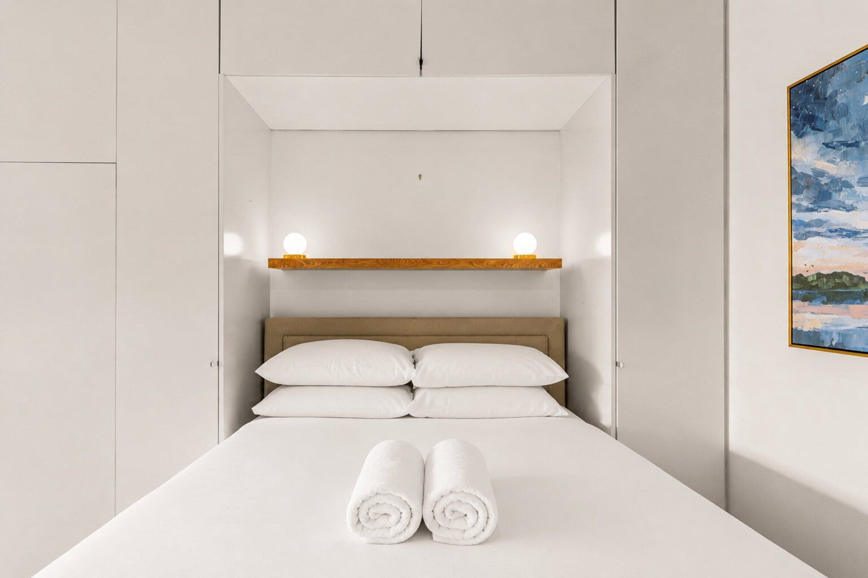 Cama bem arrumada com roupa de cama branca e toalhas dobradas, cabeceira de madeira e prateleira com duas luminárias.
