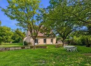 Uma casa com jardim, um pátio com cadeiras e uma árvore em primeiro plano.