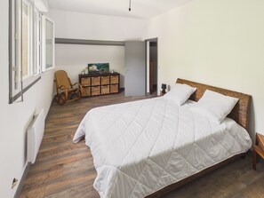 Ein Schlafzimmer mit einem großen Bett, Holzboden, einem Korbstuhl, einem kleinen Schrank und einem Bild an der Wand.