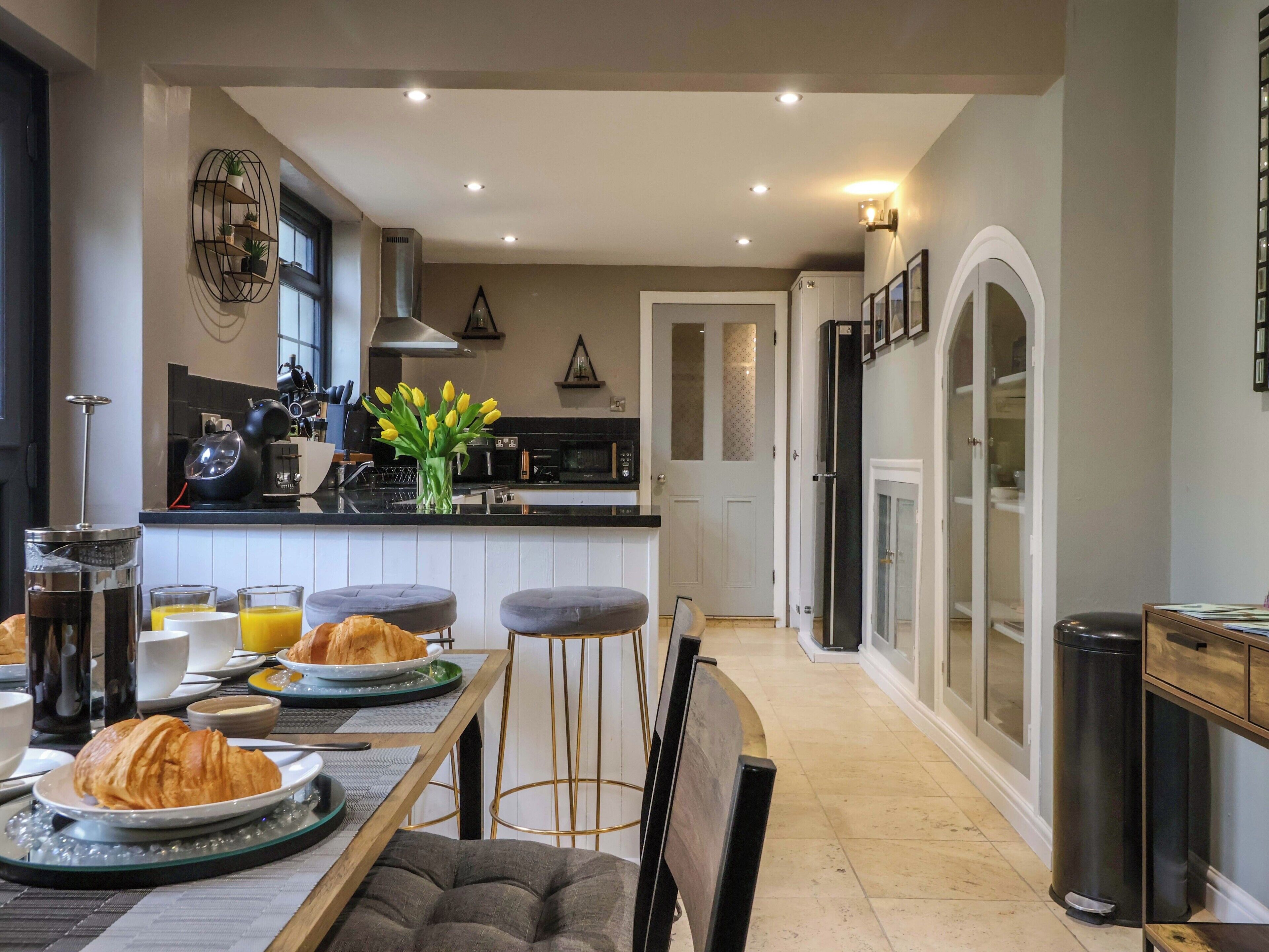 A modern kitchen with a dining area, featuring a central island, bar stools, and a breakfast setup with croissants and orange juice.