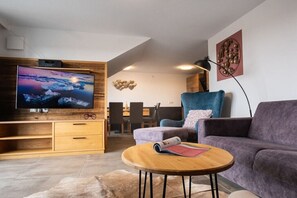 Una sala de estar moderna con televisor de pantalla plana, un mueble de entretenimiento de madera, una mesa de centro redonda, un sofá púrpura y un comedor al fondo.